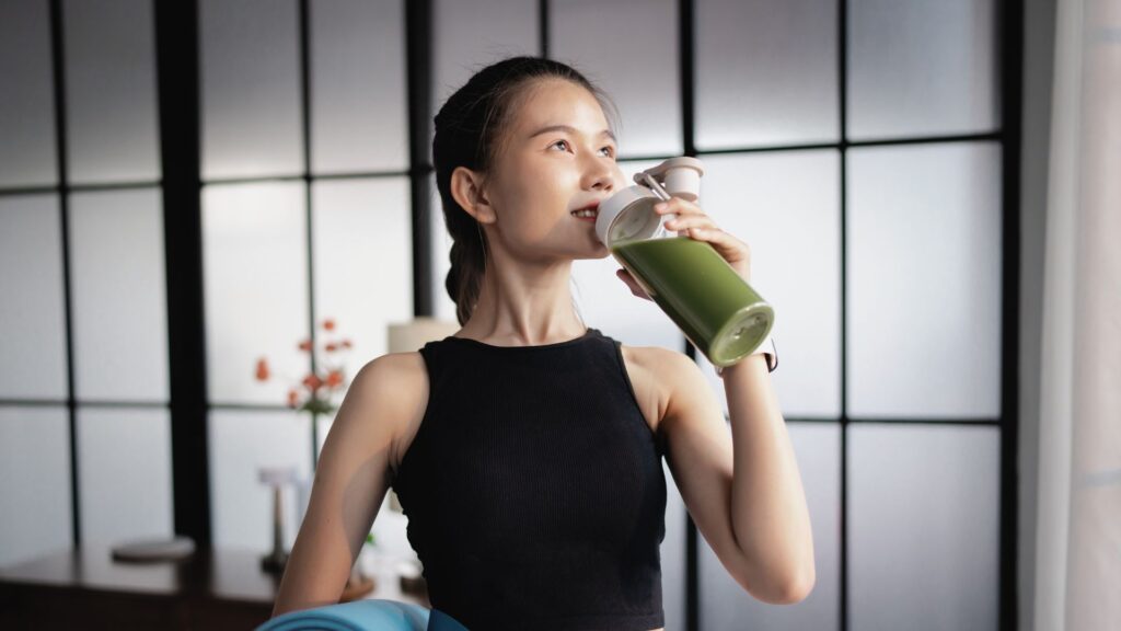 Frau mit Yogamatte und gruenem Smoothie als Symbol fuer Selbstoptimierung
