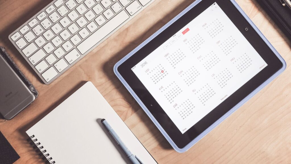 Digitaler Kalender auf Tablet neben Notizbuch und Computer