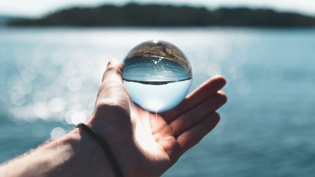 Person hält eine Glaskugel vor einem großen See - das Wasser spiegelt sich in der Glaskugel und sie sieht aus wie ein großer Tropfen